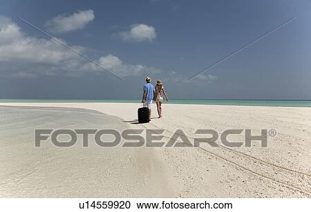 Stock Image - Couple rolling luggage on beach. Fotosearch