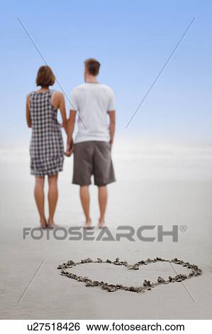 Stock Photograph - Heart in sand with couple on beach. Fotosearch