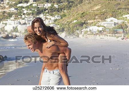 Stock Photograph - Man carrying woman piggyback on beach. Fotosearch