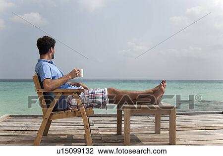 Stock Image - Man drinking coffee on deck at beach. Fotosearch