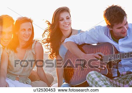 Stock Image - Man playing music for friends on beach. Fotosearch