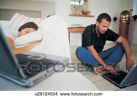 Picture - Man using laptop as girlfriend sleeps. Fotosearch