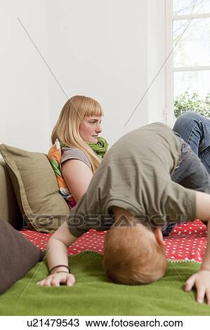 Stock Image - Mother relaxing as son jumps on sofa. Fotosearch