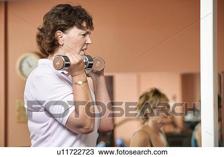 Stock Image - Older woman lifting weights in gym. Fotosearch