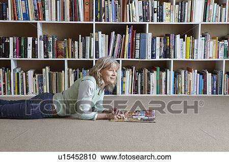 Stock Image - Older woman reading by bookshelves. Fotosearch