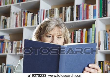 Stock Image - Older woman reading by bookshelves. Fotosearch