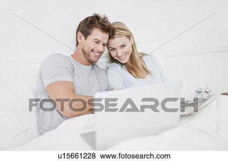 Smiling couple using laptop in bed View Large Photo Image Stock Photo - Smiling couple using laptop in bed. Fotosearch
