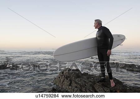 Surfer standing on rocky beach View Large Photo Image Picture - Surfer standing on rocky beach. Fotosearch