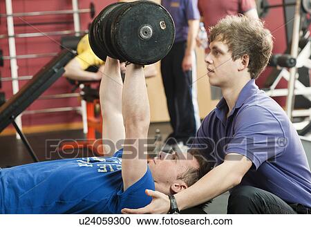 Stock Image - Trainer helping man lift weights at gym. Fotosearch