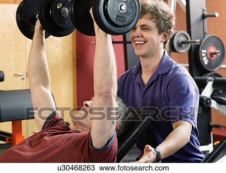 Trainer helping man lift weights at gym View Large Photo Image Stock Image - Trainer helping man lift weights at gym. Fotosearch