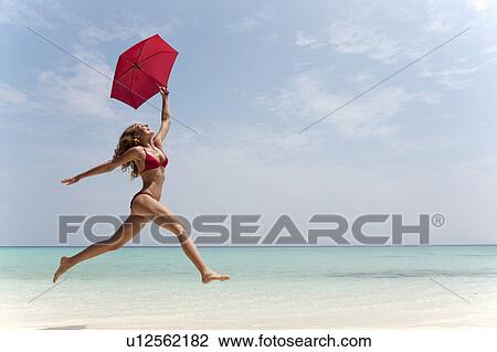 Stock Image - Woman running with umbrella on beach. Fotosearch