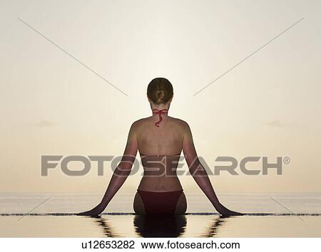 Stock Image - Woman sitting at edge of infinity pool. Fotosearch