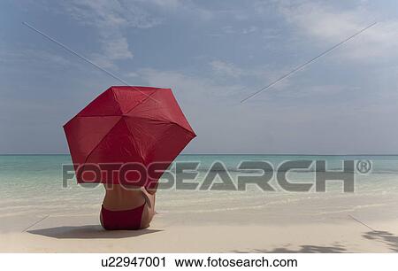 Stock Image - Woman sitting under umbrella on beach. Fotosearch