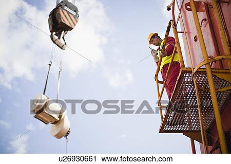 Stock Image - Worker directing crane on oil rig. Fotosearch