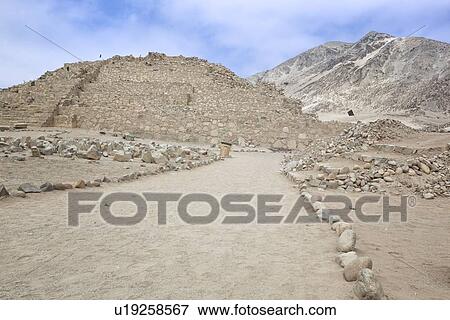 Stock Photo - Caral ancient ruins: temple mounds with mountains beyond, pale blue sky, Supe Valley, Peru. Fotosearch