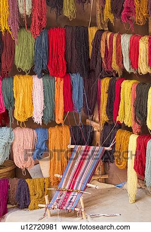 Stock Image - Close up of multi coloured skeins of wool hanging beside loom of woven Peruvian design, Awanacancha llama farm, Sacred Valley, Cuzco, Peru. Fotosearch