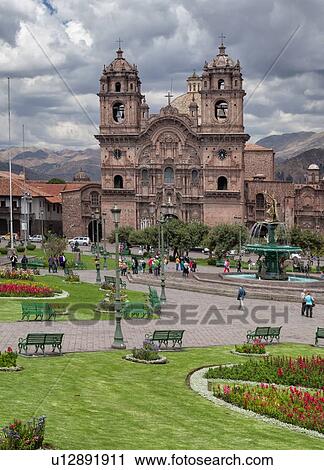 Stock Image - Cuzco: Plaza de Armas, view of Iglesia de la Compania, central fountain with statue, people walking after afternoon rain shower, blue sky with dramatic clouds, Cuzco, Sacred Valley, Peru. Fotosearch