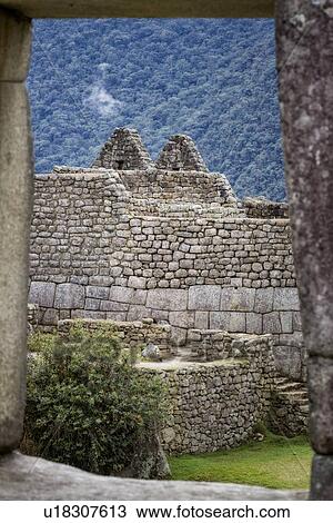Stock Image - Machu Picchu Inca ruins: detail of Eastern Urban sector terraces and buildings with steep mountain wall beyond, framed by typical Inca trapezoidal window, Machu Picchu, Sacred Valley, Peru. Fotosearch