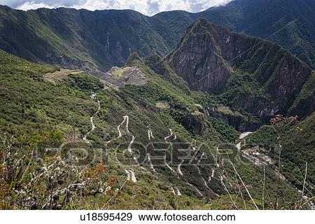 Stock Photo - Machu Picchu Inca ruins: view of entire complex: Eastern Urban sector, Royal Palaces and Temple of the Sun, Intihuatana, with Huayna Picchu and ring of mountains beyond, Hiram Bingham road snaking in foreground, as seen from Sun Gate, afternoon sunlight . Fotosearch