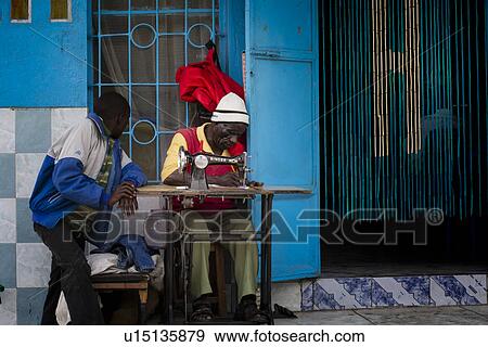 Man sitting working at sewing machine while another man leans talking, with brightly painted blue wall and beaded screen in doorway, Nakuru, Kenya, View Large Photo Image Stock Photo - Man sitting working at sewing machine while another man leans talking, with brightly painted blue wall and beaded screen in doorway, Nakuru, Kenya,. Fotosearch
