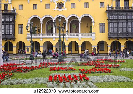 Stock Image - Plaza del Armas: detail of City Hall, yellow stucco and wooden balconies typical of Spanish Colonial era, landscaped gardens with red flowers, Lima, Peru. Fotosearch