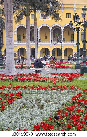 Stock Image - Plaza del Armas: detail of City Hall, yellow stucco and open balcony typical of Spanish Colonial era, landscaped gardens with red flowers, Lima, Peru. Fotosearch