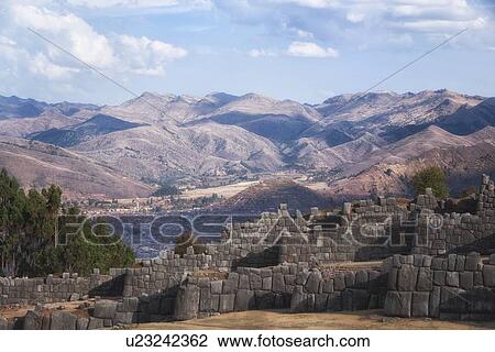 Stock Image - Sacsaywaman Inca ruins: view of the massive zig zag stone wall fortifications in late afternoon sunlight, city of Cuzco beyond, Sacred Valley, Cuzco, Peru. Fotosearch