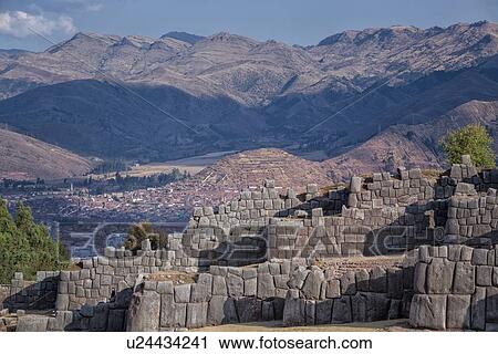 Stock Image - Sacsaywaman Inca ruins: view of the massive zig zag stone wall fortifications in late afternoon sunlight, city of Cuzco beyond, Sacred Valley, Cuzco, Peru. Fotosearch