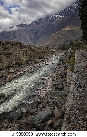 Stock Photo - Urubamba river with snow capped mountain range and dramatic clouds beyond, as seen from train from Ollantaytambo to Machu Picchu, Sacred Valley, Peru. Fotosearch