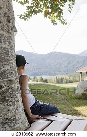 Boy sitting on bench against tree View Large Photo Image Stock Image - Boy sitting on bench against tree. Fotosearch