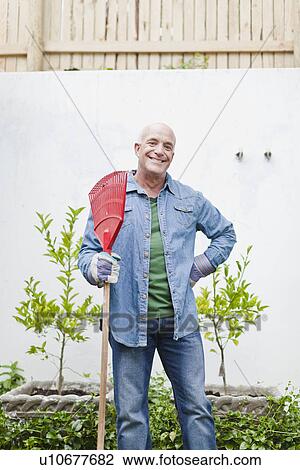 Stock Image - Older man holding rake in backyard. Fotosearch