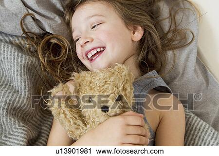 Stock Image - Smiling girl holding teddy bear in bed. Fotosearch