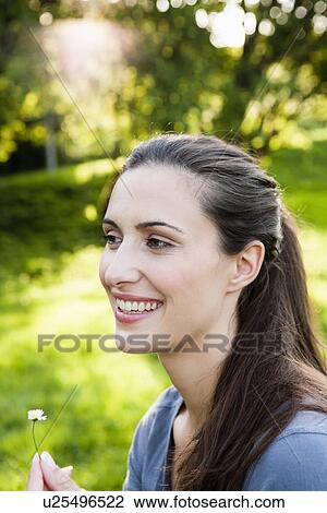 Stock Image - Smiling woman holding flower in park. Fotosearch