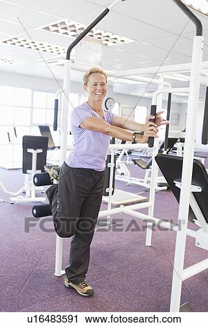 Stock Image - Woman using weights machine in gym. Fotosearch