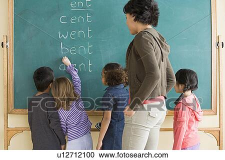 Stock Image - Children (6-7) writing on blackboard with teacher looking at them. Fotosearch
