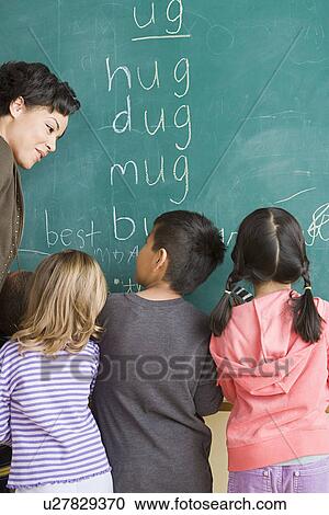 Stock Image - Children (6-7) writing on blackboard with teacher looking at them. Fotosearch