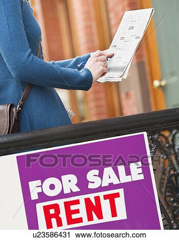 Stock Image - Close up of for sale sign with woman holding list in background. Fotosearch