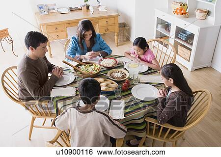 Stock Photograph - Family with three children (8-9, 10-11) praying at table before dining. Fotosearch