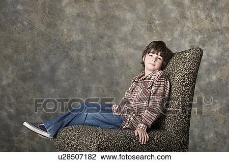 Stock Image - Portrait of boy (6-7) sitting on chair, studio shot. Fotosearch