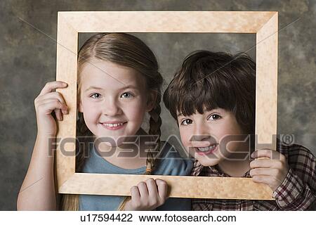 Stock Image - Portrait of girl (8-9) and boy (6-7) looking through frame, studio shot. Fotosearch