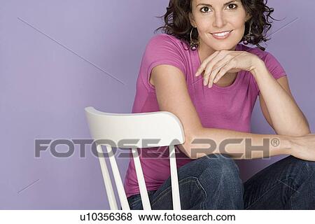 Stock Photo - Portrait of smiling woman sitting on chair with purple background, studio shot. Fotosearch