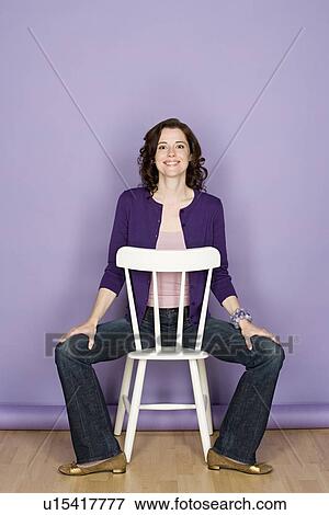 Stock Photo - Portrait of smiling woman sitting on chair with purple background, studio shot. Fotosearch