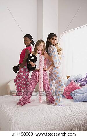Stock Image - Portrait of three girls (10-11) standing on bed at slumber party. Fotosearch