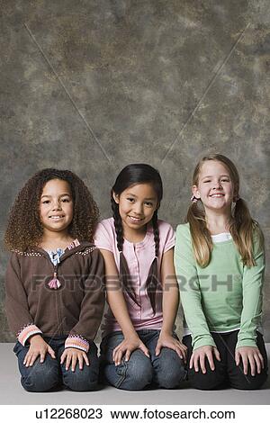 Stock Image - Portrait of three smiling girls (8-9) sitting together, studio shot. Fotosearch