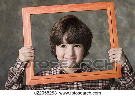 Stock Image - Smiling boy (6-7) looking through frame, studio shot. Fotosearch