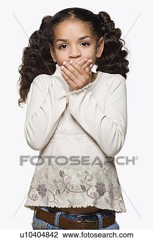 Stock Image - Studio portrait of girl (8-9) with pigtails covering mouth with hands. Fotosearch