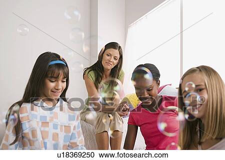 Stock Image - Three girls (10-11) and woman blowing bubbles at slumber party. Fotosearch