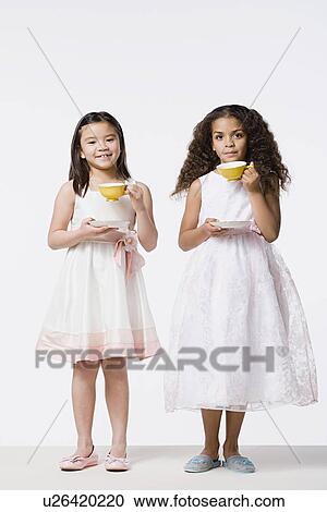 Stock Image - Two girls (8-9) wearing dresses and drinking tea, studio shot. Fotosearch