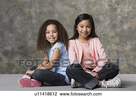 Stock Image - Two smiling girls (8-9) sitting together, studio shot. Fotosearch