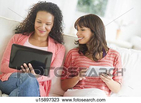 Stock Image - Two women sitting on sofa using digital tablets. Fotosearch